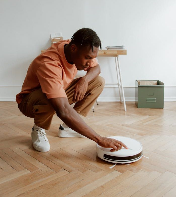 About A man kneels on a wooden floor adjusting a robotic vacuum in a minimalist home interior.
