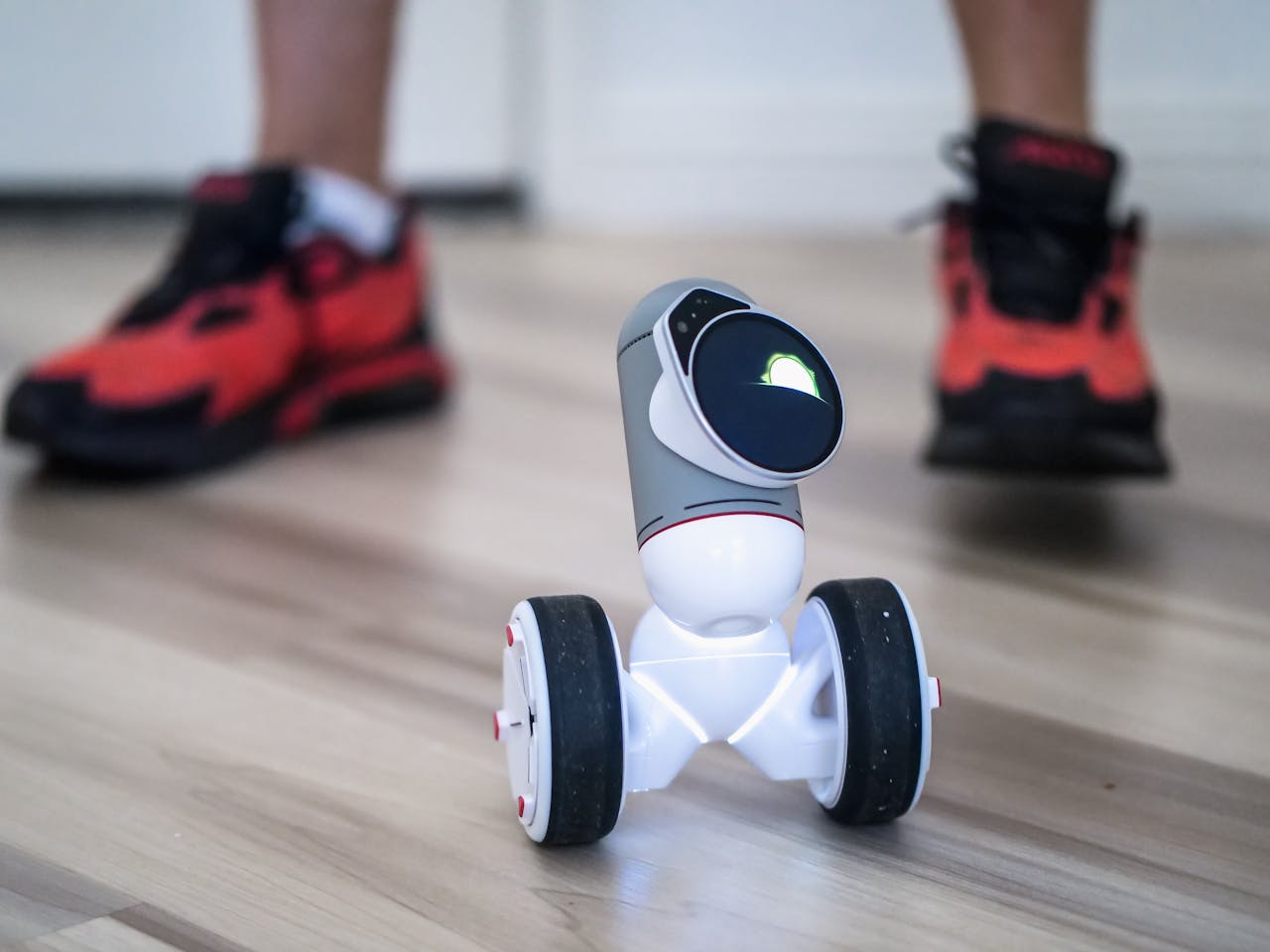 Home Close-up of a two-wheeled mini robot on a wooden floor with sneakers in background.